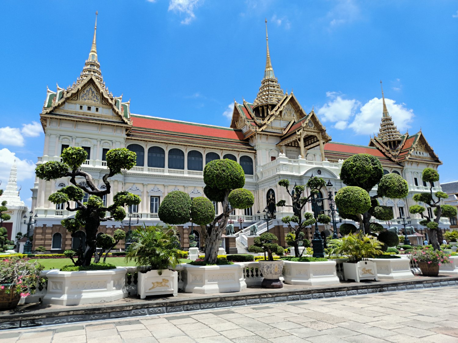 A ground view of the Grand Palace, a large complex of traditional Thai architecture, surrounded by trees and other greenery.