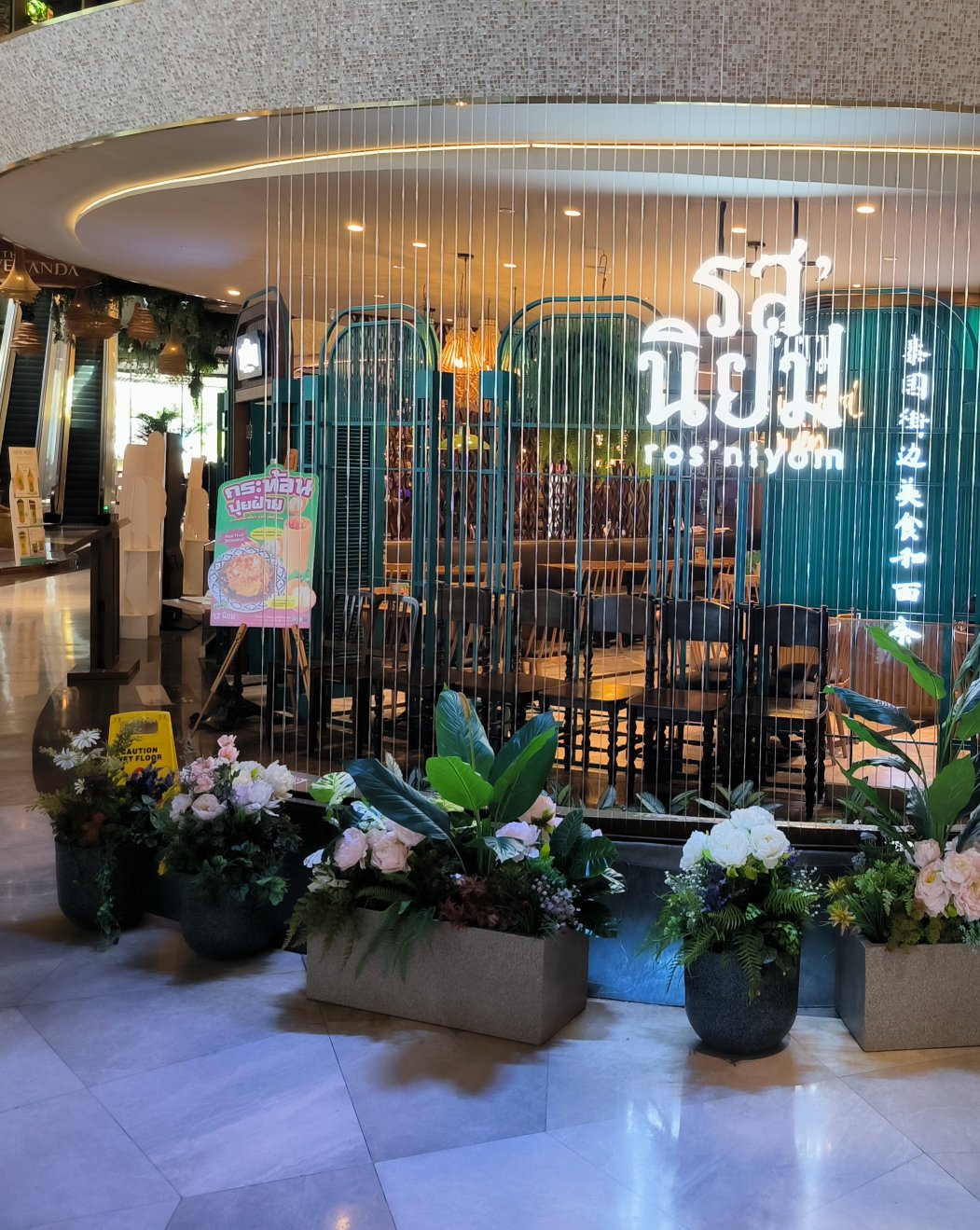 A secluded area within ICONSIAM, with a cascading waterfall resembling metal bars partially obscuring the view of a cozy restaurant named "ros niyom".